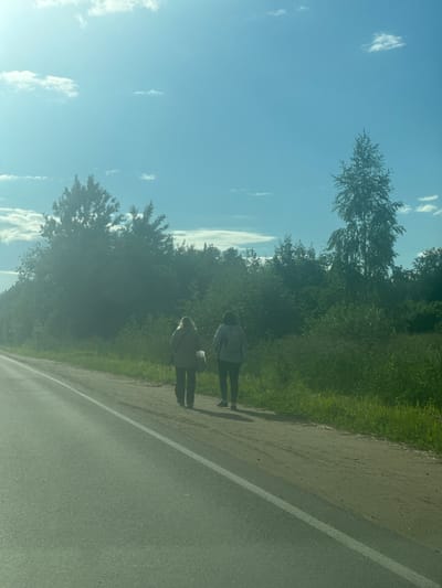 Two people are walking along the side of a paved road. One person is in front of the other. They are both wearing casual clothing. The person in front is wearing a light-colored jacket or hoodie and dark pants. The person behind is wearing a similar light-colored jacket or hoodie and dark pants. They are walking on a dirt shoulder next to the road. To the left of the road is a grassy area with trees and bushes. The sky is blue with some scattered white clouds. The sun is shining, suggesting it is daytime, likely late morning or afternoon. The overall scene appears to be a rural or semi-rural setting. There is no visible text in the image.