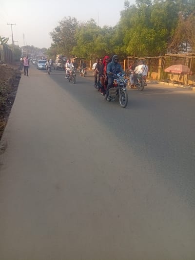 A busy street scene shows multiple motorbikes carrying passengers, including one with four individuals. Other motorbikes, cars, and a pedestrian are visible further ahead. The street is wide, and trees line the side, behind a fence. A red umbrella with “4G” is present near the right edge. Overall, the image depicts a typical urban environment in a developing country.