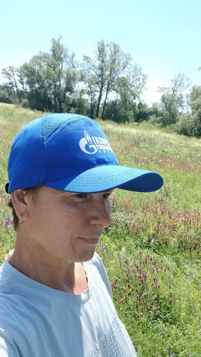A woman is seen from the chest up, facing right, against a backdrop of a sunny field with trees and a clear sky. She is wearing a blue baseball cap with a white logo that appears to be a gas company, and a light blue t-shirt. She has short hair and is wearing a small hoop earring in her left ear. The field is covered in green grass and scattered purple and yellow wildflowers. The scene suggests an outdoor activity in a rural or semi-rural setting. The location is specified as Nadezhdenka, Russia.