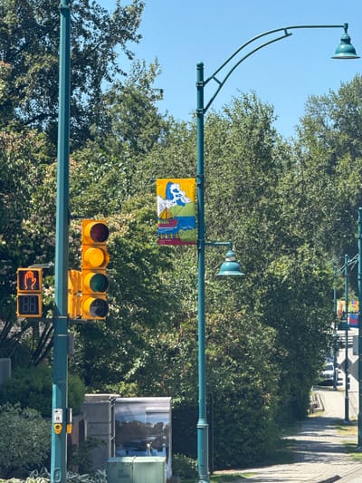 The image shows a street scene in Port Moody, Canada, on a clear, sunny day. A teal colored lamppost stands prominently on the left, displaying a traffic light with a red hand symbol indicating "Don't Walk" above a digital display showing "88". Below the traffic light, a pedestrian crossing button is visible on the lamppost. To the right of the lamppost, another teal lamppost supports a street lamp and a decorative banner featuring a boat on water and a blue and white cloud formation, with text "CORA WANG 2023" at the bottom. The street is lined with lush green trees and foliage. A paved sidewalk runs along the right side of the street, leading into the distance where more lampposts and banners are visible. A white vehicle is parked further down the street on the right. In the foreground on the left, a grey utility box with a reflective surface is partially visible, mirroring the sky and trees.
