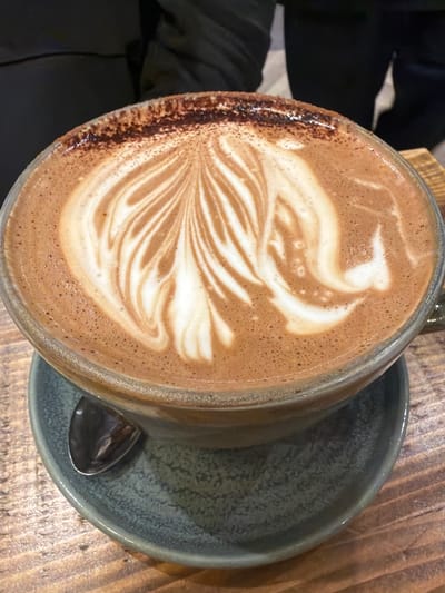 A close-up, angled shot focuses on a cup of latte art resting on a blue-grey saucer placed on a wooden surface. Part of a black-clad individual is visible to the left. The coffee cup is filled with a beverage topped with an intricate white pattern resembling a leaf or feather design. The cup's rim appears dusted with cocoa powder. A small spoon sits in the saucer. The environment suggests a cafe or restaurant setting, possibly in Mole Valley, United Kingdom.