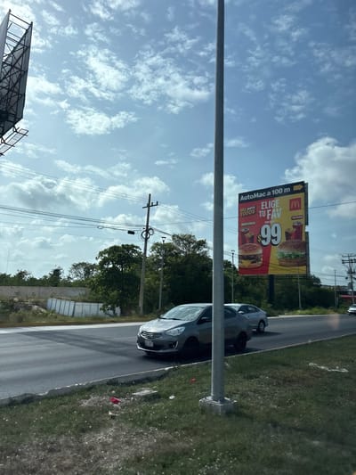 The image shows a highway with a billboard in the background. The billboard is for McDonald's and advertises a promotion for 99 pesos. There are two cars on the highway, and the sky is cloudy. The location is Cancún, Mexico. The billboard is located at kilometer 100 of the AutoMac. The billboard is illuminated, and it is daytime. The billboard is advertising two burgers for 99 pesos, one is a BBQ Cheddar burger and the other is a McPollo burger. There are also drinks displayed on the billboard. The highway is a multi-lane road with a concrete barrier. There are trees and vegetation on the side of the road. There is a street light and power lines in the image. There is also a large metal structure on the left side of the image, which could be part of a larger advertising structure or a construction site.