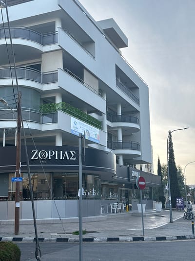 The image presents a modern urban street scene in Agios Dometios, Cyprus, under an overcast sky during daytime. The primary subject is a multi-story building featuring a clean, contemporary design with light grey facades, numerous windows, and curved glass balconies. Some balconies are adorned with lush greenery.

On the ground floor of this building, a commercial establishment is visible with a large, prominent sign displaying "ZORPAS 1975" in white lettering on a black background. An outdoor seating area with several white chairs and tables is set up on the paved sidewalk in front of this establishment, suggesting a café or restaurant.

In the foreground and midground, a street runs alongside the building, bordered by distinct black and white checkered curbs. A red circular 'do not enter' traffic sign is clearly visible near the street. High above the outdoor seating, a bicycle is uniquely positioned or mounted on a pole, adding an unusual detail to the streetscape. Utility poles and overhead power lines crisscross the lower portion of the image.

On one of the building's balconies, a banner is displayed with Greek text, including "MEΛETHTHPIO ΦΡΟΝΤΙΣΤΗΡΙΟ" (Study/Tutoring Center) and "Solidus," indicating the presence of educational or professional services on the upper floors. Further down, a small blue street sign is partially visible, though its text is illegible.

No people are visible in the scene, contributing to a quiet and still atmosphere. The weather appears mild, with no harsh shadows, consistent with an overcast day. The overall setting is that of a well-maintained commercial and residential area.