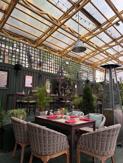 This is a medium-angle, eye-level, exterior shot of an outdoor dining area in Zhukovka, Russia. The scene is set under a translucent, corrugated roof supported by a yellow wooden frame, adorned with string lights. The dining area features dark wooden tables and chairs, some with woven rattan seating. Each table is set with red napkins, glassware, and what appear to be salt and pepper shakers. Lush green plants, including potted ferns and a cypress tree, are strategically placed around the seating areas, adding to the ambiance. A tall, pyramid-shaped patio heater stands to the right of the frame. In the background, a dark green wooden fence is visible, decorated with framed pictures and a decorative clock-like structure with potted plants. The overall impression is that of a well-appointed, comfortable outdoor dining space, likely a restaurant patio.