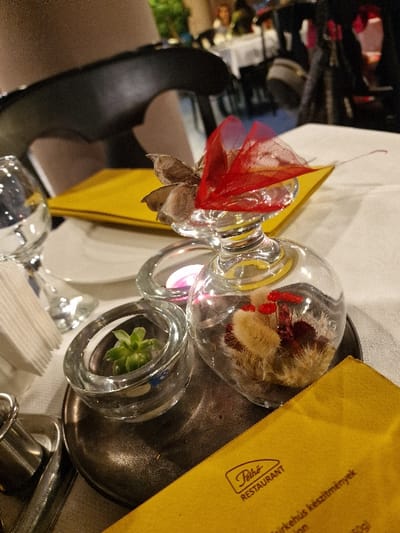 This image depicts a table setting inside Pethő Restaurant in Odorheiu Secuiesc, Romania. The foreground features a white tablecloth upon which a circular, dark metallic tray is positioned. On this tray, a collection of decorative glass items is arranged. A central heart-shaped glass ornament contains dried plant material, including fluffy white and red elements, and is topped with several brown, seed-like pods and a translucent red fabric bow. To its left, a small cylindrical glass holder contains a green succulent plant. Adjacent to that, another similar glass holder contains a lit pink votive candle.
A yellow menu cover, bearing the "Pethő Restaurant" logo and Hungarian text, rests diagonally on the tray and tablecloth. Partially visible objects on the table include a white ceramic plate, a stemmed glass (possibly for wine or water), a stack of white paper napkins, and a silver utensil. The background is out of focus, showing the interior of the restaurant with other tables set with white tablecloths, dark chairs, and blurred figures of individuals, suggesting other patrons. The lighting is dim and warm, consistent with an evening dining atmosphere.