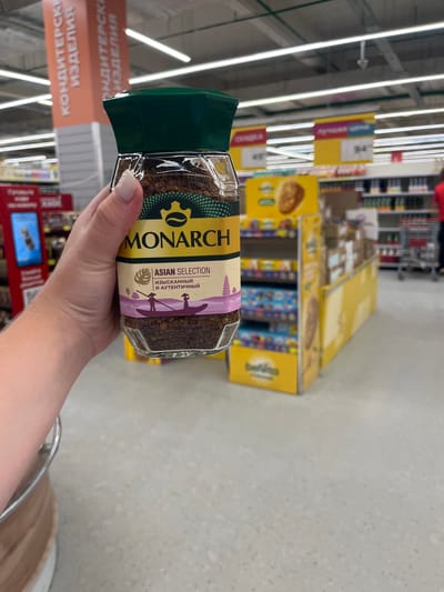 A person's hand, with painted fingernails, holds a glass jar of Monarch brand instant coffee. The jar is labeled in English and Russian. The background shows a retail environment, likely a supermarket in Volzhsky, Russia, with aisles of products, promotional displays, and overhead lighting. A sign with Cyrillic text, possibly indicating "Confectionery Products," is visible in the upper left. A price tag with "94" is displayed on a yellow promotional stand. The floor is a light-colored speckled material.
