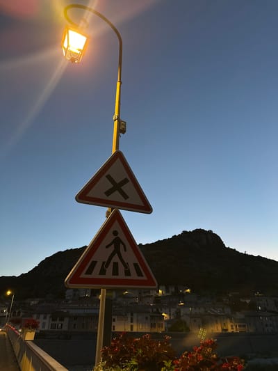 The image depicts a street scene in Anduze, France, at dusk. Two triangular road signs are prominent: the upper one displays a black "X" indicating an intersection, while the lower one shows a silhouette of a pedestrian crossing a crosswalk. Both signs have red and white borders. They are mounted on a yellow pole, which also supports a street lamp emitting a warm, yellow light. The sky is a clear, deep blue, transitioning from lighter shades near the horizon. In the background, a dark, tree-covered mountain looms over a village. The village's buildings are visible, with some lights illuminated in windows. The foreground features a railing, possibly part of a bridge or walkway, with planters containing red flowers. The overall atmosphere is serene, marking the transition from day to night.