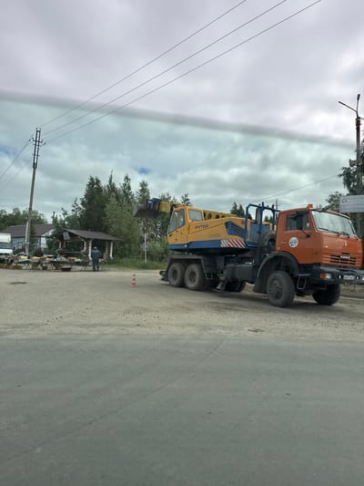 An overcast day frames a scene in Parma, Russia, where a large orange Kamaz truck, equipped with a yellow crane attachment labeled "АНТЕЙ," is parked on a dirt lot. The crane arm is raised. To the left of the truck, a small market stall is set up under a wooden gazebo, with several tables displaying goods. Two individuals, presumably vendors or shoppers, are present near the stalls. A single orange traffic cone is visible in the foreground, between the market and the truck. Power lines stretch across the sky, supported by utility poles. The overall environment suggests a rural or semi-urban setting, possibly on the outskirts of the city.