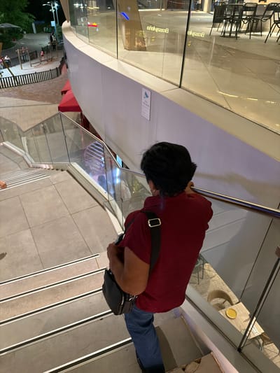 The image shows a person walking down a staircase in what appears to be an indoor shopping mall or a similar public building. The person is wearing a dark red long-sleeved shirt and blue jeans and has a black bag slung across their chest. They are looking down towards the stairs as they descend.
The staircase has a modern design with a metal railing and tiled steps. The background shows a reflection in glass surfaces, with glimpses of seating areas and architectural features that suggest a commercial or public space. The lighting suggests it is evening or nighttime, as there are reflections of artificial lights.
The scene is set in Alcorcón, Spain. There are no clear indications of the weather, but the indoor setting implies it is not a factor for the activity shown. The mood is neutral, with no strong emotional cues from the subject. No visible text is discernable in the image.