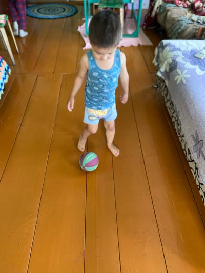 A young child wearing a blue tank top and light blue shorts stands on a wooden floor, looking down at a small, multi-colored ball at their feet. The ball is primarily green and purple. The child's left foot is positioned slightly in front of their right. The floorboards are a warm, orangey-brown color and appear to be well-worn. In the background, a patterned blanket is draped over a piece of furniture on the right side of the frame, and a round rug with concentric circles is visible on the floor to the left. The overall setting suggests a domestic indoor environment.