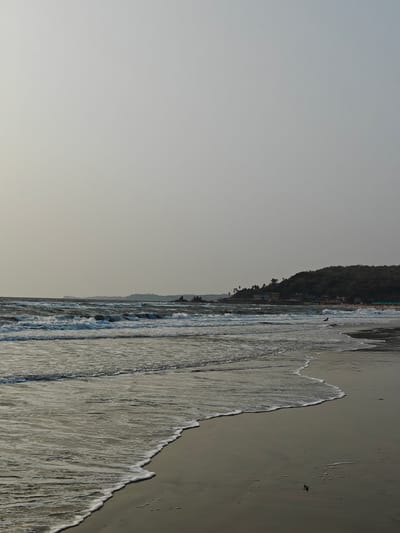 The image depicts a shoreline in Arambol, India. Waves with white foam crests roll onto a dark sand beach under an overcast sky. In the distance, a headland topped with vegetation is visible, with some buildings nestled along the coast. People are present on the beach to the right of the headland and one person is in the water.