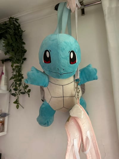 An indoor scene in Saint-Leu-la-Forêt, France, depicts a blue Squirtle plush backpack, featuring a cream-colored patterned belly and red eyes, hanging from its light blue fabric straps over a chrome-finished metallic rod. A light pink bag, adorned with a white floral pattern and white straps, is suspended below the Squirtle via a white strap and silver-colored carabiner clip. Additional light-colored fabric items are draped over the metallic rod. The metallic rod is affixed to a white wall. To the right, a white curtain hangs from the rod. To the left, a section of a white shelf holding various containers is visible below artificial green foliage cascading against the white wall.