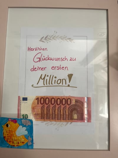 The image shows a framed piece of paper with a handwritten message in German that reads "Herzlichen Glückwunsch zu deiner ersten Million!" which translates to "Congratulations on your first million!". Below the text, there are several stylized images of banknotes. To the left of the banknotes, there is a child's drawing of a yellow chicken with a pink bow on its head and a gray cloud in a blue sky. The framed item is displayed in what appears to be a domestic setting, with a pale pink frame. The image does not contain information about the location in the city of Schwäbisch Hall, Germany.