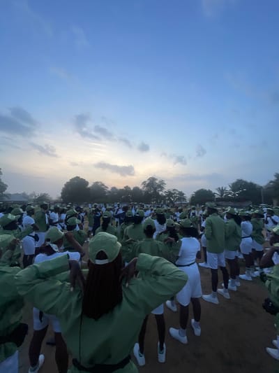 In the city of Ise-Ekiti, Nigeria, a large group of young adults, predominantly women, are gathered outdoors for what appears to be a morning assembly or orientation. They are all dressed in matching khaki uniforms consisting of green shirts, white shorts, and green caps, with white socks and white sneakers. The scene is captured at dawn or dusk, with the sky displaying soft hues of blue and scattered clouds, illuminated by the rising or setting sun.
The individuals are arranged in a field, with a scattering of trees visible in the background, suggesting a rural or semi-urban setting. Some are standing in rows, while others are in more informal clusters, facing in various directions. The general atmosphere appears to be one of order and collective activity, possibly related to a national youth service or a similar program. The low lighting indicates it's either early morning or late evening, contributing to the diffused, soft quality of the light. The ground is a dirt or gravel surface.