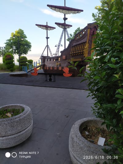 An outdoor shot captures a playground designed to resemble a pirate ship, located in Avcılar, Turkey. The ship, constructed of wood with painted details and fabric sails, is positioned on a black rubberized surface. In front of the ship, two orange elephant-shaped spring riders are visible. The ground leading to the playground is paved with gray tiles, and two large, concrete planters filled with vegetation are in the foreground. To the right, lush green foliage partially obscures the scene. The sky above is a clear blue with a few wispy clouds. The image is timestamped June 18, 2026, at 06:36.
