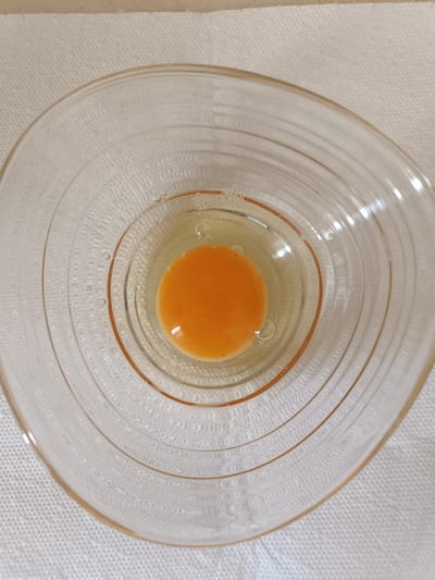 An overhead, close-up shot shows a single raw egg yolk and white in a clear, glass bowl. The bowl has a rippled, concentric design on its interior surface and is placed on a white, textured paper napkin. The background is a light beige surface. There are no people or animals visible in the image. The location is not specified and cannot be determined from the image.