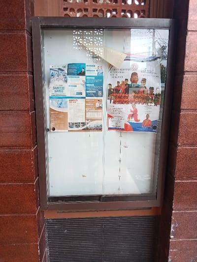 The image shows a bulletin board located on a brick wall. The bulletin board is made of wood and glass and has two sections. The left section has a poster that reads "Santri Baru" and displays images of people in various settings. The right section has a poster that reads "Latihan Bersama" and displays images of people in a martial arts setting. The background is a brick wall and the location is Kecamatan Brangsong, Indonesia.