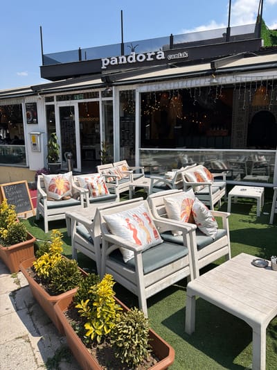 The image shows the outdoor seating area of a restaurant called "Pandora Çamlık" in Denizli, Turkey. White wooden chairs and tables are arranged on a patch of artificial grass, with several potted plants in brown planters flanking the seating area. Decorative pillows with Egyptian-themed designs adorn the chairs. In the background, the restaurant facade is visible, featuring large glass windows and doors, with the "Pandora" logo prominently displayed. The sky is clear and blue.