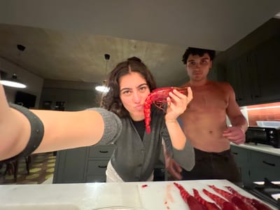 A photograph taken with the camera turned upside down shows two people, a woman and a man, presumably in a kitchen. The woman is wearing a gray cardigan over a white undershirt and holding a bright red shellfish, possibly lobster, in her hands. She is looking at the camera. The man, who is shirtless, stands beside her. A counter or countertop is visible in the lower half of the image. There are other pieces of what appear to be shellfish parts on a white surface in the background. The location is identified as Gondarém, Portugal, based on the available context. Kitchen cabinetry and appliances are visible in the background, with hanging light fixtures above.