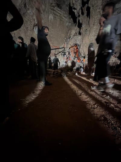 The image depicts a group of people inside a dimly lit cave, likely a tourist attraction, with the ground covered in sand and rocks. In the background, the cave walls are rough and textured, with stalactites hanging from the ceiling. Several individuals are visible, some standing and others sitting, appearing to observe or engage with the cave's features. The lighting is low, casting long shadows across the ground, with some areas illuminated by artificial light sources, possibly from flashlights or the ambient light of the cave's setup. There are also what appear to be decorative elements or religious figurines placed within the cave, some of which are illuminated with an orange glow. The overall atmosphere suggests a guided tour or a visit to a natural wonder. It is not possible to confirm the specific location as Guttapadu, India, based solely on the visual information provided in the image.