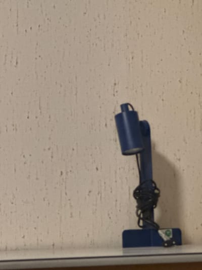 The image captures a wall-mounted blue fixture in what appears to be an indoor or covered outdoor setting in Schömberg, Germany. The fixture consists of a multi-jointed arm, extending horizontally from the right side of the frame. At the end of the arm is a cylindrical, dark blue element, possibly a light or sensor. A bundle of tangled black electrical wires is coiled around the central part of the blue arm. The fixture is attached to a light beige or off-white wall, characterized by a textured surface resembling concrete or stucco with numerous small, dark brown flecks. Near the wall-mounting point on the blue fixture, a small sticker or label is visible with a green, roughly circular logo, but no discernible text due to the image's blur. No people are present, and no specific activity is taking place. The scene is static, under what appears to be artificial or dim lighting.