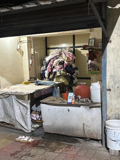 This image depicts the open-fronted interior of a modest, ground-level establishment, likely a laundry or ironing shop, located in Ambernath, India, during evening or night hours. A dark, corrugated metal shutter is partially rolled up overhead.

The interior is dimly lit. To the left, a light-colored wall shows cracks and stains, with an exposed electrical switchboard and wires. A large, rectangular ironing table, covered with a light-colored fabric or tarp, occupies the left-center. A pair of blue jeans is laid on the table, along with a yellow rectangular object. Below the table, a white tarp is draped, concealing items, with a grey bag visible underneath.

Against the back wall, a substantial, unorganized pile of various colorful garments, including pinks, blues, and whites, is stacked high, indicating a bulk laundry operation. A small, dark-colored animal, possibly a dog or cat, is partially visible at the base of this pile, nestled behind a counter.

To the right of the clothes pile, a dark metal frame is visible. Further right, a shelf holds a framed picture, a red bag-like object, a QR code poster, and a hanging plastic bag containing items. A green and white poster with Hindi text is also affixed to the wall.

In the right-center, a roughly constructed counter with peeling paint or plaster supports several items: a plastic water bottle, an orange plastic bucket, a red terracotta water pot (matka) with a tap on a stand, and a white ribbed plastic container. A blue QR code payment stand (Paytm UPI) and a set of keys are also on this counter.

The floor in the foreground is composed of reddish and greyish interlocking paver blocks, appearing damp in areas. A pair of light-colored sandals with straps rests on the pavers to the left. To the far right, a white plastic bucket with blue markings sits on a small brick block next to a blue-grey concrete pillar that frames the right side of the opening. No human individuals are visible in the scene.