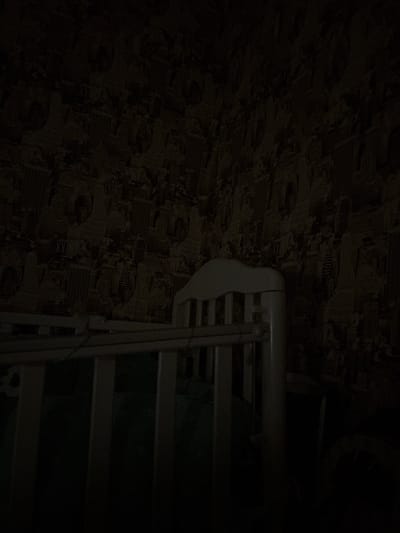 The image shows an indoor scene, possibly a bedroom or nursery, with a crib in the foreground. The crib is white with vertical slats and a curved headboard. The walls are covered in patterned wallpaper depicting a cityscape with skyscrapers and text that appears to be in English, including "CITY," "HOME," and "LIGHTHOUSE." The lighting is dim, casting long shadows and making it difficult to discern finer details of the room. There are no people or active events depicted in the image. The location is specified as Sovkhoza "Razdol'e", Russia, however, there is no visual evidence within the image to confirm this specific geographical location; the wallpaper design with English text suggests a possible non-Russian context or a deliberate thematic choice for the decor.
