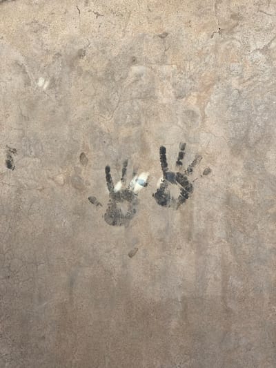 A close-up vertical image depicts a textured, light grey-tan concrete surface. Centrally positioned are two distinct human handprints, rendered in a dark, likely carbonaceous or pigmentary, residue. Both handprints are oriented vertically with fingers pointing upwards and are situated side-by-side. The residue coverage within the prints is somewhat irregular, showing variations in opacity. The concrete surface exhibits minor hairline cracks and subtle tonal inconsistencies across its visible area. This scene is located in Kokran, Afghanistan.