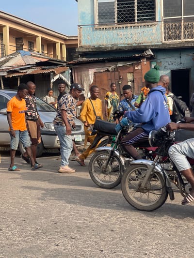 The image depicts a street scene in Akowonjo, Nigeria, with several individuals and motorcycles. In the foreground, a man in a blue hoodie and green beanie is seated on a motorcycle, facing right. Behind him, another motorcycle with a rider is partially visible on the right. To the left, a group of men are walking along the street. One man in an orange t-shirt and denim shorts is at the front of this group, followed by a man in a patterned shirt and brown shorts, and another man in a patterned shirt and jeans. A silver car is parked behind them. In the background, there are buildings, including one with a blue facade and a balcony with ornate railings. Signs are visible on the buildings, one reading "THIS HOUSE is NOT 4 SALE". The overall environment appears to be an urban setting with weathered infrastructure.