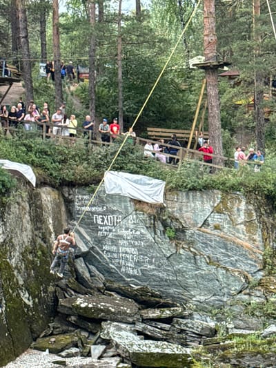 In the city of Chemal, Russia, a man is ziplining down a rocky cliff face. He is wearing a brown t-shirt and dark pants, with a harness for the zipline. The zipline is a yellow rope. The cliff is covered in inscriptions, some of which are visible in the image. The cliff is surrounded by trees and lush green vegetation. In the background, a group of people are watching the zipliner from a wooden platform. The platform is elevated and has a wooden railing. The overall scene depicts an outdoor adventure activity taking place in a natural, mountainous environment.