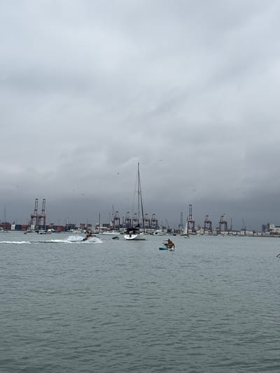 An overcast day at the port of Callao, specifically La Punta, Peru, features a body of water with several vessels and a distant industrial skyline. In the foreground, a person is operating a jet ski, creating a spray of water. Further out, a person is stand-up paddleboarding on a blue board. A white sailboat with a black sail is anchored to the right. In the background, numerous cargo ships and large cranes are visible, indicating a busy port. Seagulls are scattered across the sky, and the water is choppy with a gray hue.