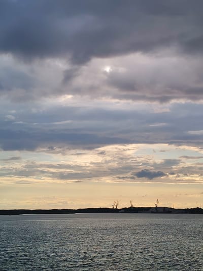 The image depicts a seascape with a harbor in the distance. The sky is filled with dramatic clouds, with patches of sunlight breaking through. The water is choppy, reflecting the overcast sky. In the background, along the shoreline, a dark silhouette of trees is visible, with a few industrial cranes and structures rising above them. There are no people visible in the image. The overall impression is one of a working harbor under a moody sky.