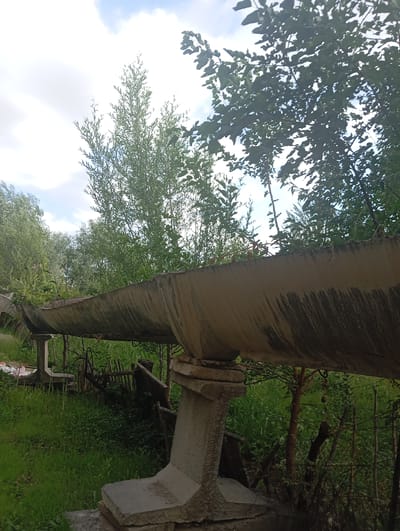 The image shows a close-up of a weathered, curved concrete structure, possibly an aqueduct or a similar infrastructure element, supported by concrete pillars. The structure is set against a backdrop of lush green foliage and trees. The sky above is partly cloudy with patches of blue. The ground is covered in grass and dense vegetation, with a rustic wooden fence visible in the middle ground. The overall scene suggests an outdoor, possibly rural or semi-urban, environment. The image does not depict any people or specific actions. The location in the city of Kızıl-Bel, Kyrgyzstan is not visually discernible from the image itself.
