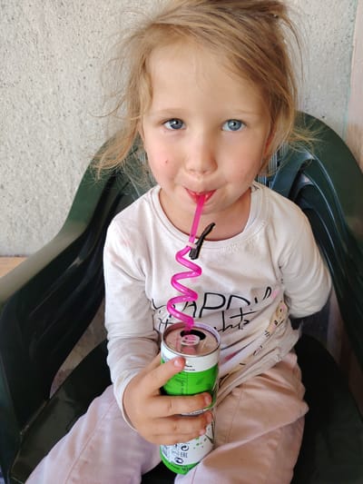 A young girl with blonde hair and blue eyes is seated in a green plastic chair, drinking from a green and white can with a pink curly straw. She is wearing a light-colored long-sleeved shirt and light pink pants. The background is a textured, light gray wall. The location appears to be outdoors, possibly in Byala Slatina, Bulgaria.