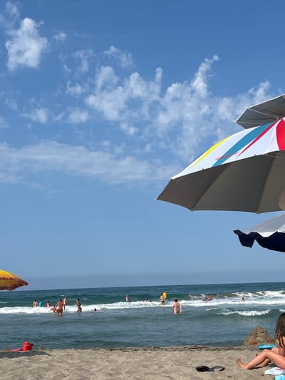 This media file captures a lively beach scene under a clear blue sky with scattered white clouds. The perspective is looking upwards, with the edge of a colorful beach umbrella dominating the top left portion of the frame. The umbrella displays segments of red, white, yellow, and blue.
Below the umbrella, the ocean stretches out, with gentle waves rolling towards the sandy shore. Several people are visible in the water, enjoying the sea, some wading and others swimming. The beach itself is a sandy expanse, with a few scattered objects and people visible on it. One person's legs and feet are prominently featured in the top right, suggesting they are sitting on the sand. The time of day appears to be daytime, likely late morning or early afternoon, given the bright sunlight and clear skies, indicative of good weather. The overall mood is one of leisure and recreation. There is no visible text in the image. The location is Ahtopol, Bulgaria.