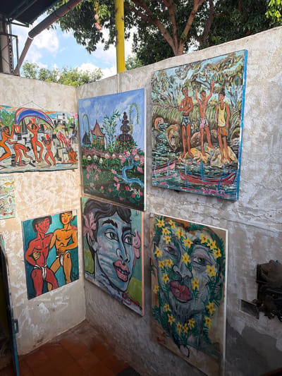 The image depicts an outdoor art display against a concrete wall in Tejakula, Indonesia. Several paintings are mounted on the wall. In the upper left, a large horizontal painting features figures engaged in what appears to be a harvest or ritualistic activity, with elements resembling buildings and water. To its right, a vertical painting shows a landscape with a pagoda-like structure, lush greenery, and pink flowers, with a flowing blue ribbon element. Above this, another vertical painting displays three young men standing near water, with boats visible below. In the lower left corner, a square painting portrays two bare-chested men in dynamic poses. To the right of that, a large vertical painting dominates the scene, featuring a close-up of a person's face rendered in blue tones, adorned with pink flowers and green foliage. Below this, another vertical painting presents a person's face covered in yellow flowers and green leaves. The environment includes rough concrete walls, a tiled floor in the foreground, overhead foliage from trees, and a glimpse of the sky with clouds. The overall impression is of an artist's outdoor studio or a small exhibition space in a tropical setting.