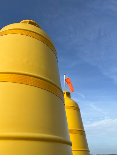The image captures a low-angle view of two large, yellow cylindrical objects against a bright blue sky with scattered white clouds. These objects appear to be industrial tanks, possibly for storage, and are banded with reflective yellow tape at intervals. One of the tanks has an orange windsock attached to its upper section, indicating it might be an outdoor installation exposed to the elements. The sky suggests it is daytime, likely with clear weather given the brightness. The perspective is unusual, looking upwards towards the sky, emphasizing the scale of the tanks. There is no visible text in the image. The scene is likely an industrial area or an open landscape in Terhillion, Ireland, given the contextual information.
