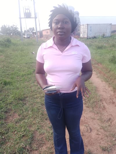 A medium shot photo shows a Black woman standing outdoors. She is wearing a pink polo shirt with the logo "alo" in white, dark blue jeans, and a black and white headwrap. She is holding a small white object, possibly a phone case, in her left hand. The background features a grassy area, a dirt path, and a distant view of a power transformer, buildings, and a shipping container. The image is likely taken in Agwala, Nigeria, based on the provided context.