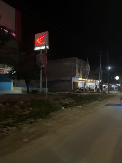 This is a nighttime, outdoor, low-angle shot of a street in Bundar, Indonesia. The scene depicts a Honda dealership. In the foreground, a brightly lit Honda sign with its distinctive wing logo illuminates the dark. To the left, a portion of a building is visible, with some areas appearing to be under renovation or construction. The building's facade is a muted beige, contrasting with the vibrant red of the Honda logo.
Behind the dealership, a street stretches into the distance, lined with more buildings and illuminated by streetlights and the light spilling from the businesses. Several motorcycles are parked in front of the dealership, suggesting it is either closing or has recently closed for the day. Some figures, likely customers or staff, are visible inside the brightly lit establishment, engaged in conversation or activity. A red and white banner hangs from a flagpole in front of the dealership, with visible text that appears to be in Indonesian, though it is partially obscured by the banner's folds and the angle of the shot. One section of text clearly reads "HONDA." The overall atmosphere is one of a typical Indonesian street scene at night, with the buzz of commerce present despite the late hour. The road surface is visible, appearing to be paved and slightly damp, reflecting the ambient light. The presence of utility poles and wires indicates that the area is well-connected.