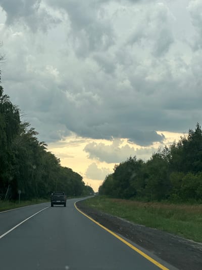 The image depicts a highway scene in Pereyezd, Russia, with a dark-colored SUV driving on the right side of the road. The road has a yellow line on the right edge and a white line in the center. Dense green trees line both sides of the road, forming a tunnel-like effect. The sky is dramatic, filled with heavy, grey clouds, but a bright, golden light breaks through them on the left side, suggesting either sunrise or sunset. The overall mood is somber due to the stormy clouds, yet hopeful due to the light breaking through. The time of day appears to be either early morning or late evening. The activity is simply driving on a highway, with the SUV in the foreground indicating movement. No people are clearly visible, and no text is discernible on the SUV or any other objects.
