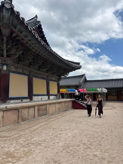 Two women walk across a sandy courtyard in front of a traditional Korean temple building in Gyeongju-si, South Korea. The temple features a dark, tiled roof with ornate eaves and colorful painted decorations. Behind the temple, another building with a similar tiled roof is visible, adorned with strings of colorful lanterns. The sky above is partly cloudy with patches of blue. The woman on the left wears a white tank top and dark pants, carrying a brown shoulder bag. The woman on the right wears a black tank top and dark, flowing pants, holding a black umbrella. Several other people are seen walking up stairs in the background, near the lanterns. The ground is covered in light brown sand, with a low stone wall bordering the temple building.