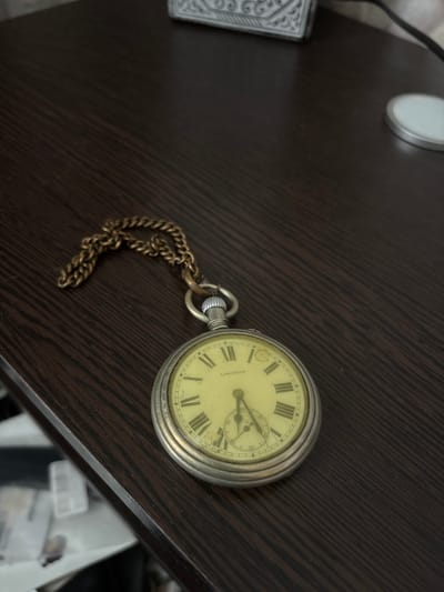 A close-up, high-angle shot shows a vintage Longines pocket watch with a gold-colored chain resting on a dark brown wooden surface. The pocket watch has a cream-colored face with black Roman numerals and a small sub-dial at the six o'clock position. The watch chain is looped around the watch. The background is out of focus, but suggests a desk or table with other objects, including a decorative white object on the upper left and a round silver object on the upper right. The image does not contain any elements that can identify the location as Kinel'-Cherkasy, Russia.