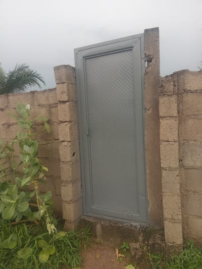 The media file shows a gray, diamond-patterned metal door set into a rough-hewn brick wall. The door is rectangular and appears to be a hatch or a small entrance. The brickwork surrounding the door is uneven and appears weathered, with some sections crumbling. To the right of the wall, there is lush green grass and foliage, indicating an outdoor setting. A palm frond is visible in the lower left corner, suggesting a tropical or subtropical environment. The sky is overcast and appears a pale gray. The overall impression is of a functional, utilitarian structure in an undeveloped or rural area. There is no text visible in the media file. The image is oriented sideways. The context provided indicates the location is Gbaza, Nigeria. The time of day appears to be daytime, but the overcast sky does not provide specific clues.