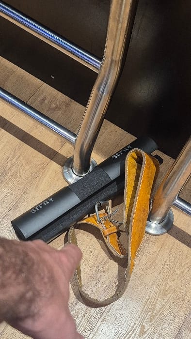A black Hilus barbell pad and leather weightlifting belt rest atop a light-colored wooden floor. The barbell pad is cylindrical with a textured center. The leather belt has a yellow suede-like interior and a tan exterior with a metal buckle and loop. The items sit near the chrome legs of a weight bench. A hand and forearm reach into the scene on the right edge.