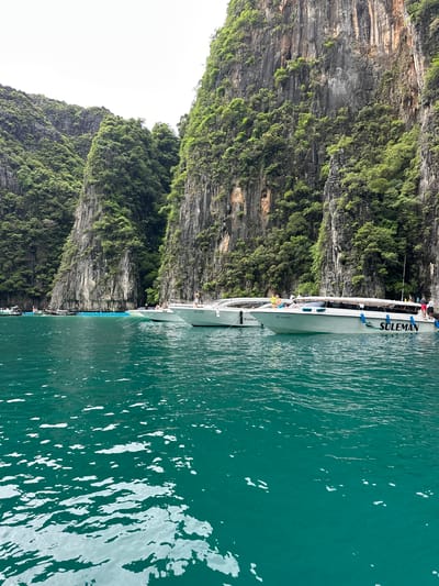 A group of longtail boats are anchored in the turquoise waters off the coast of Haad Yao, Thailand. The boats are white with blue trim and have the names "LOVE ARC" and "SULEMAN" written on their sides. People can be seen standing on the decks of the boats and swimming in the water. The background is dominated by towering limestone cliffs covered in lush green vegetation. The sky is overcast.