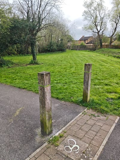 This image depicts a grassy area in Swindon, United Kingdom. The foreground features a paved path with a cycle lane symbol painted on the brickwork. Two wooden posts with green bands are positioned on either side of the path, marking an entrance to the grassy area. The grass is well-maintained with visible mowing lines. In the background, a wooden bridge spans a ditch or stream, with houses visible beyond the bridge. The trees are in early leaf, suggesting springtime. The sky is overcast. There are no people visible in the image.