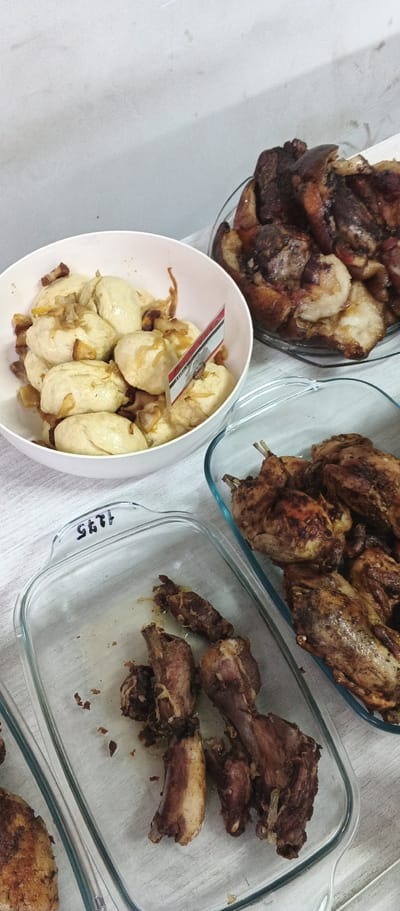 The image shows a table laden with food, likely a meal being prepared or served. In the foreground, two glass baking dishes contain cooked meat. The dish on the left holds several pieces of what appear to be ribs or small cuts of pork, some with visible bone. The dish on the right is filled with what seem to be small, whole roasted birds, possibly quail or Cornish game hens, which are browned and appear to be cooked. In the background, a white bowl contains several dumplings or stuffed buns, topped with what looks like sautéed onions and pieces of meat, possibly bacon. A small flag with red, white, and black stripes is stuck into the dumplings. To the right of the bowl, a larger glass dish is filled with more pieces of cooked meat. The food is arranged on a light-colored surface, which could be a table or countertop, with a white background behind it. There are no people visible in the image. The presence of the flag suggests a potential national or regional context for the meal, but no specific location details such as "Ploske, Ukraine" are discernible from the visual evidence.