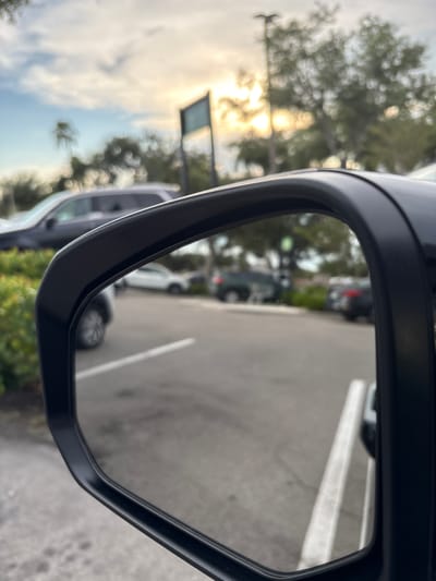 The image shows a rear-view mirror of a car parked in a lot, with a view of other cars and trees reflected in the mirror. The sky is cloudy with sunlight breaking through. The scene appears to be in South Venice, United States of America. There are several cars visible in the reflection, including a dark SUV, a white SUV, and a dark sedan. The environment is a parking lot with some greenery and trees in the background. No people are visible in the image.