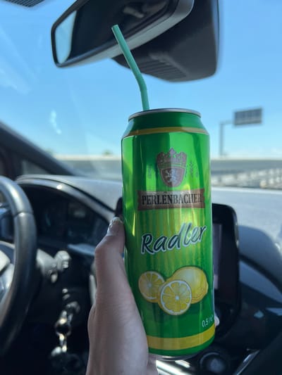 A hand with painted fingernails holds a green can of "PERLENBACHER Radler" with a green straw inserted into it. The can is positioned in the foreground, with the driver's side dashboard and steering wheel visible in the background. The scene appears to be inside a car, with a rearview mirror and a bright sky visible through the windshield. The image does not contain any visual information or context related to the city of Barletta, Italy.