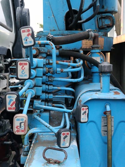 The image shows a close-up view of a blue hydraulic control panel, likely from a piece of heavy machinery. The panel is covered in various levers, tubes, and connectors, many of which are rusted. Several of the levers have small, square labels with red and white pictograms and Japanese text. One label shows an arrow pointing up and to the right with the text "伸" (extend), and another shows arrows pointing down and to the left with the text "縮" (retract). Another label features a hook symbol with "上" (up) and "下" (down) indicated. A beige-colored lever is also visible with Japanese text "左" (left) and "右" (right) and a curved arrow. There is a metal hose attached to the panel with a clamp. The background is slightly blurred and suggests an outdoor environment with some green foliage. The scene is located in Ban Patan, Thailand, which implies it is an industrial or construction setting.